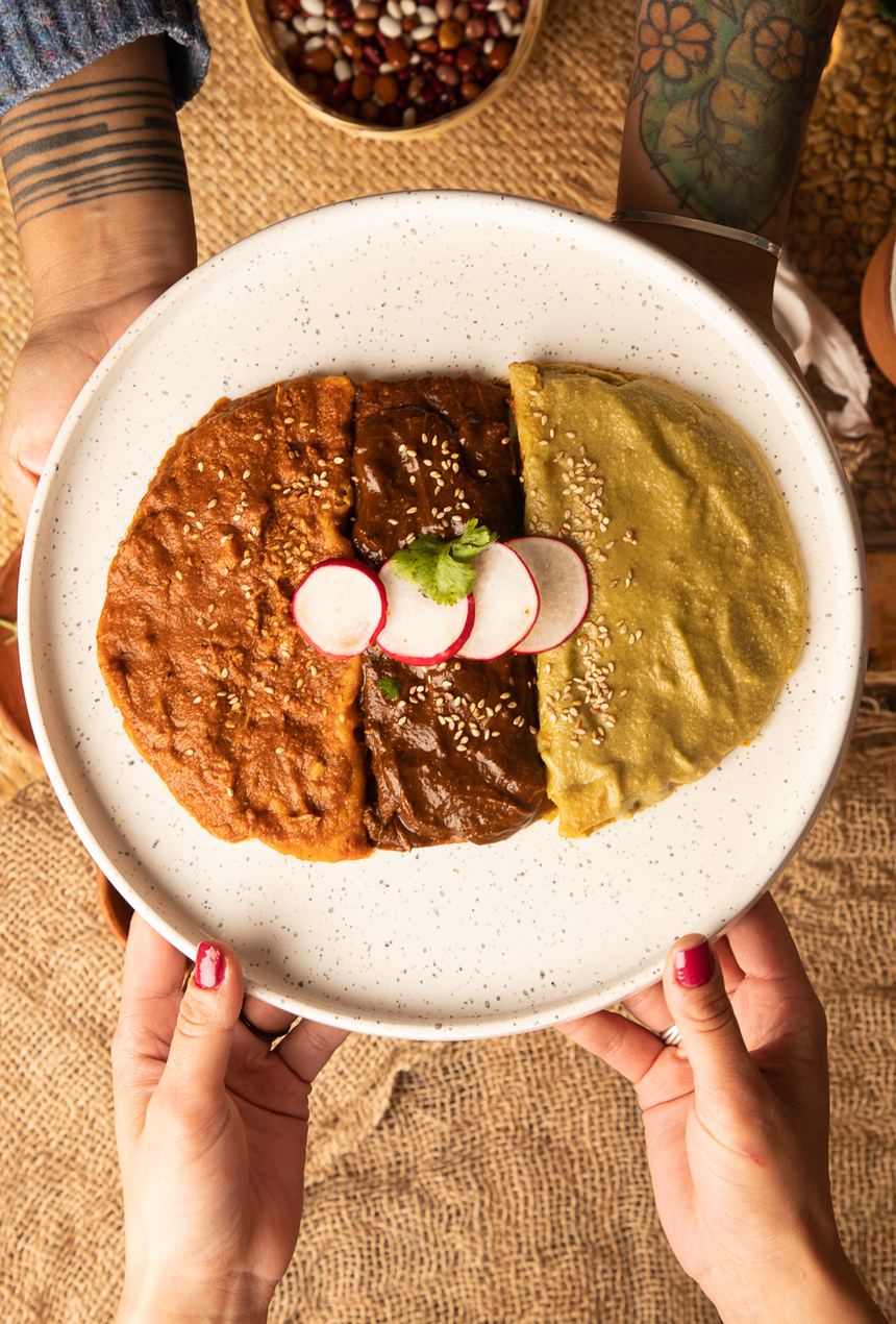 Dos pares de manos sostienen un plato de enmoladas de pipián rojo, mole y pipián verde