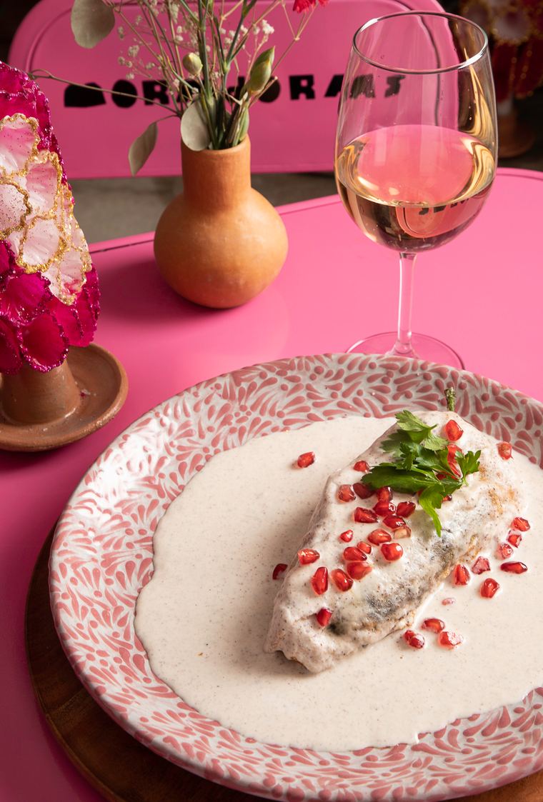 Un chile en nogada servido sobre una mesa rosa, junto a una copa de vino y flores
