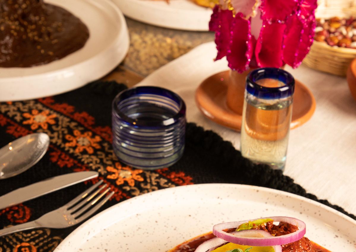 Una mesa servida en el comedor: el mole, las chalupas, la chancla, flores de papel y vasos de vidrio soplado sobre un mantel bordado