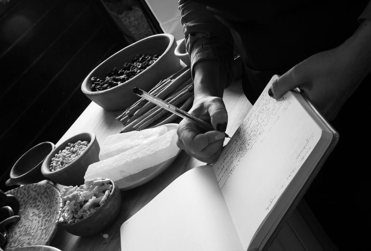 Una mano escribe la receta en un cuaderno, entre cajetes de pasas, canela, piñón y queso